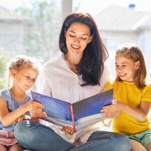 Uma mulher sorridente está sentada lendo um livro para duas meninas pequenas sentadas ao seu lado.
