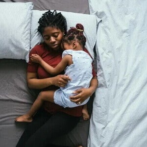 Mãe solo – os desafios das mulheres que criam filhos sozinhas Foto de uma mãe solo dormindo com a filha no colo em sua cama. As duas estão no meio da cama que está com as laterais vazias