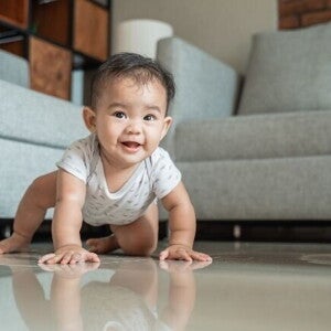 Um bebê sorridente engatinhando em um piso brilhante, com um sofá cinza e uma parede de tijolos ao fundo, ilustrando um marco de desenvolvimento motor. Um bebê sorridente engatinhando em um piso brilhante, com um sofá cinza e uma parede de tijolos ao fundo, ilustrando um marco de desenvolvimento motor.