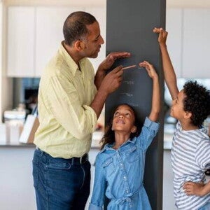 Em uma cozinha, está um homem, uma menina e um menino. A menina está encostada em uma parede, enquanto o homem e o menino marcam na parede qual é a altura dela. Em uma cozinha, está um homem, uma menina e um menino. A menina está encostada em uma parede, enquanto o homem e o menino marcam na parede qual é a altura dela.