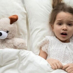 Uma menina assustada está deitada na cama, coberta por um lençol, com expressão de medo, ao lado de um cachorro de pelúcia. Uma menina assustada está deitada na cama, coberta por um lençol, com expressão de medo, ao lado de um cachorro de pelúcia.