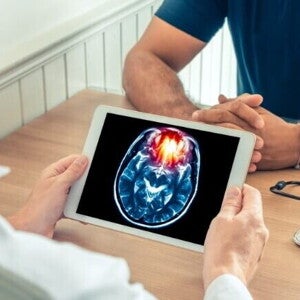 Um médico de jaleco branco mostra um tablet com uma imagem de ressonância magnética de um cérebro para um paciente. A imagem ilustra as graves consequências de doenças como a meningite B.