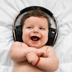 Um bebê sorridente, deitado em uma superfície branca, usa fones de ouvido grandes e pretos sobre a cabeça.