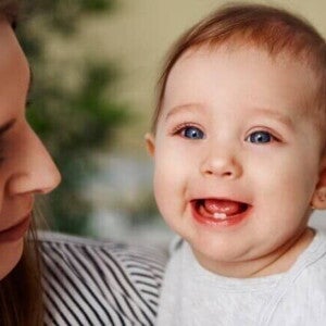 Bebê sorrindo no colo da mãe, com os primeiros dentinhos visíveis, ilustrando o processo do nascimento dos dentes do bebê.