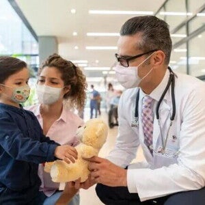 Neuropediatra Neuropediatra e enfermeira se aproximam de uma criança, que segura um urso de pelúcia.