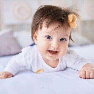 Bebê sorridente deitada de bruços na cama, usando roupa branca e um laço no cabelo Bebê sorridente deitada de bruços na cama, usando roupa branca e um laço no cabelo