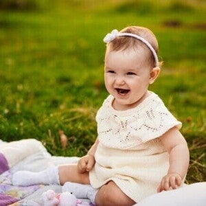 Bebê sorridente usando um vestido claro e uma tiara com flores, sentada em um cobertor sobre a grama. Bebê sorridente usando um vestido claro e uma tiara com flores, sentada em um cobertor sobre a grama.