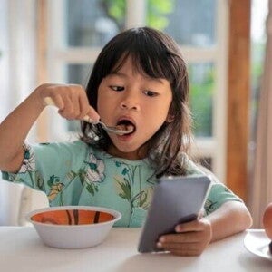 Uma menina come enquanto olha para um celular na mesa. Uma menina come enquanto olha para um celular na mesa.