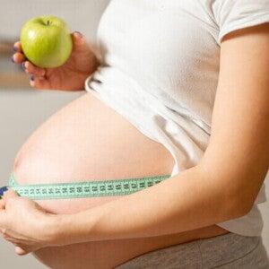 Mulher grávida segurando uma maçã em uma mão e uma fita métrica para costura na outra, ao redor da barriga. Mulher grávida segurando uma maçã em uma mão e uma fita métrica para costura na outra, ao redor da barriga.