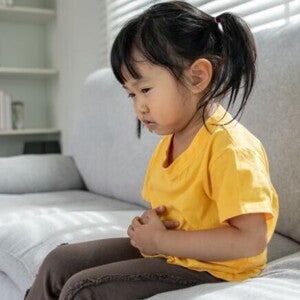 Uma menina está sentada em um sofá, segurando a barriga com as duas mãos, com uma expressão de desconforto ou dor. Uma menina está sentada em um sofá, segurando a barriga com as duas mãos, com uma expressão de desconforto ou dor.
