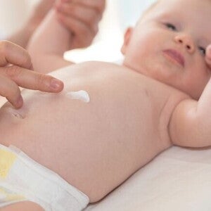 Uma mão de adulto gentilmente aplica creme na barriga de um bebê deitado, destacando o cuidado com a pele delicada do bebê. Uma mão de adulto gentilmente aplica creme na barriga de um bebê deitado, destacando o cuidado com a pele delicada do bebê.