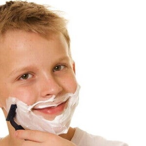 Um menino passando com creme de barbear no rosto, passando uma lâmina de barbear na bochecha e sorrindo Um menino passando com creme de barbear no rosto, passando uma lâmina de barbear na bochecha e sorrindo