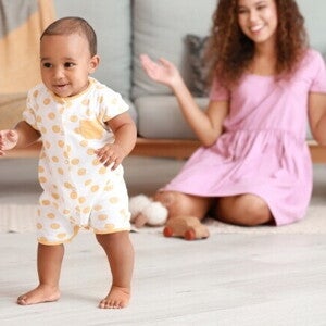 Um bebê sorrindo e em pé no chão, olhando para a frente, com uma mulher sentada no chão ao fundo, sorrindo.