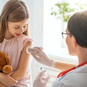 Uma menina segura um ursinho de pelúcia enquanto uma profissional de saúde, prepara o braço da menina para uma injeção com um algodão.