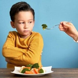 Um menino de blusa amarela expressa recusa alimentar infantil ao cruzar os braços e desviar o olhar de um garfo com brócolis oferecido por um adulto. À sua frente, há um prato com legumes cozidos sobre uma mesa de madeira e fundo azul.