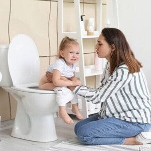 Mãe consolando uma criança pequena que chora e está sentada no vaso sanitário, ilustrando o desconforto e preocupação associados a problemas intestinais ou a presença de sangue nas fezes de criança.