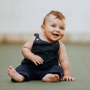 Bebê pequeno, de bochechas rechonchudas e olhos azuis, sentado em um chão plano e sorrindo, vestindo um macacão jeans azul-marinho.