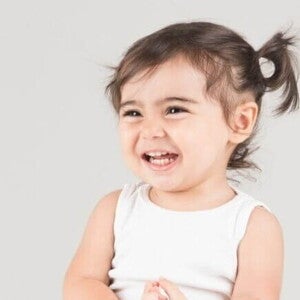 Close-up de um bebê sorrindo, com o cabelo preso em um rabo de cavalo no topo, vestindo uma regata branca, com fundo cinza-claro.