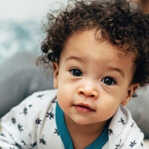 Bebê com cabelo cacheado vestindo um pijama branco com detalhes azuis, olhando diretamente para a câmera. Bebê com cabelo cacheado vestindo um pijama branco com detalhes azuis, olhando diretamente para a câmera.