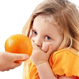 Uma mão oferece uma laranja a uma menina, que cobre a boca com a mão e olha para a câmera com uma expressão de repulsa ou aversão.