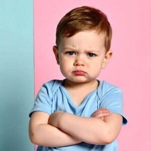 Fotografia de um menino pequeno expressando teimosia infantil. Ele está de braços cruzados, com uma expressão facial fechada e lábios comprimidos, vestindo uma camiseta azul contra um fundo dividido entre azul claro e rosa.