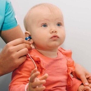 Profissional da saúde aplicando o teste da orelhinha em um bebê no colo, usando um pequeno fone no ouvido da criança para verificar a capacidade auditiva.