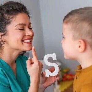 Uma mulher sorrindo, apontando para a própria boca e segurando um objeto em forma de letra “S”, enquanto uma criança observa e tenta imitar a ação da mulher. Uma mulher sorrindo, apontando para a própria boca e segurando um objeto em forma de letra “S”, enquanto uma criança observa e tenta imitar a ação da mulher.