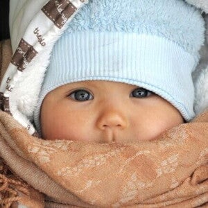 Um close-up de um bebê usando um chapéu azul e um cobertor marrom enrolado em volta do rosto, mostrando como agasalhar um bebê para o frio e como vestir um bebê de acordo com a temperatura.