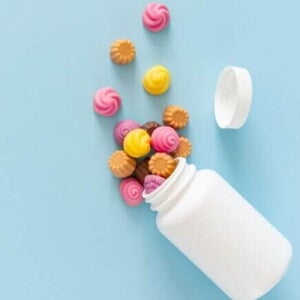 Visão de cima de gomas de vitaminas coloridas, em formato de espiral ou flor, que caíram de um pote branco sem rótulo, sobre uma superfície azul clara.