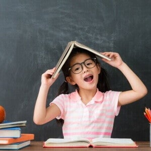 Uma menina com óculos, sentada à mesa, faz uma pose divertida com um livro aberto sobre a cabeça. Ela sorri com a boca aberta em frente a um quadro negro, com um monte de livros e lápis de cor ao seu lado. A imagem é lúdica e representa a criatividade e a diversão do aprendizado, remetendo ao clima de volta às aulas. Uma menina com óculos, sentada à mesa, faz uma pose divertida com um livro aberto sobre a cabeça. Ela sorri com a boca aberta em frente a um quadro negro, com um monte de livros e lápis de cor ao seu lado. A imagem é lúdica e representa a criatividade e a diversão do aprendizado, remetendo ao clima de volta às aulas.