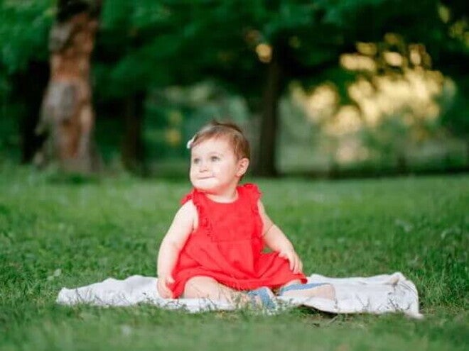 Sua criança completou um ano? Preparar, explorar, descobrir! Criança de um ano com vestido vermelho e fita no cabelo posa para foto em um parque, sentado em um pequeno cobertor colocado sobre a grama.