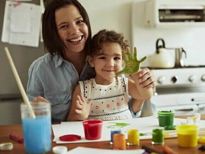 Sentadas à mesa estão uma menina e uma mulher. Espalhadas pela superfície temos tintas de diversas cores e papéis. A mão da criança está toda pintada de verde e as duas estão alegres. A imagem remete a brincadeiras para fazer com crianças mais crescidinhas Sentadas à mesa estão uma menina e uma mulher. Espalhadas pela superfície temos tintas de diversas cores e papéis. A mão da criança está toda pintada de verde e as duas estão alegres. A imagem remete a brincadeiras para fazer com crianças mais crescidinhas