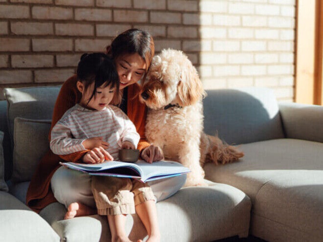 Pessoa lendo um livro ilustrado para uma criança enquanto estão sentadas no sofá com o cachorro da família