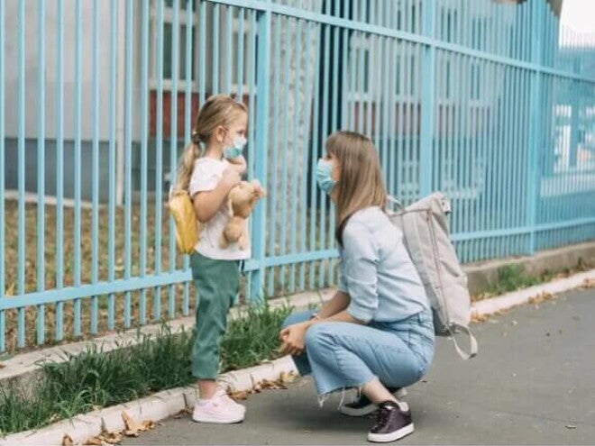 Adaptação Escolar Uma mãe está agachada em frente a sua filha, perto de um portão. A menina está de mochila, olhando para a mãe e segurando um urso de pelúcia.