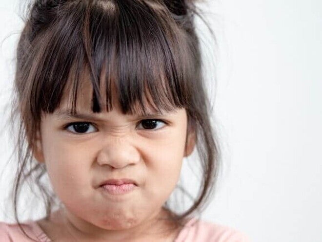 Close do rosto de uma menina expressando agressividade na primeira infância. Ela está com as sobrancelhas franzidas, nariz contraído e lábios pressionados em um sinal nítido de raiva ou frustração. O fundo é neutro e claro, destacando a expressão emocional da criança.
