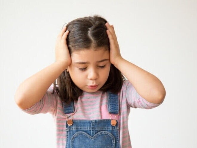 Menina de cabelo castanho com as mãos na cabeça, em uma expressão de desconforto ou dor. A imagem ilustra o sintoma, destacando a necessidade de saber como aliviar dor de cabeça em criança.
