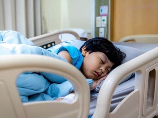 Uma criança dormindo profundamente em uma cama de hospital, enrolada em lençóis azuis, em um ambiente hospitalar.