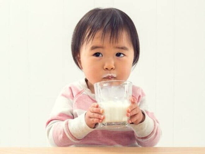 Bebê sentado à mesa, bebendo leite de um copo de vidro com bigode de leite no lábio superior. O fundo é branco.