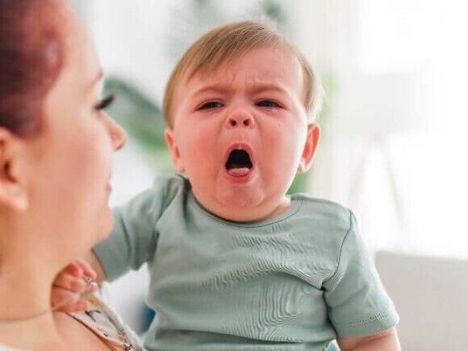 Bebê sendo segurado por uma mulher. O bebê está com a boca aberta e uma expressão de choro ou tosse.