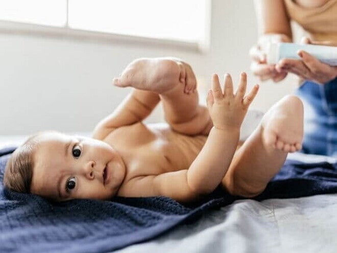 Assadura alergia alimentar Um bebê deitado de fralda em um trocador sobre uma toalha azul-escura, com as pernas levantadas. Na parte superior direita, mãos desfocadas de um adulto seguram um tubo de creme, preparando-se para aplicar pomada para assadura por alergia alimentar ou de contato.