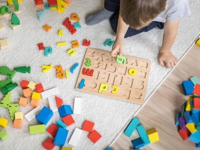 Em um piso de madeira com um tapete cinza, está sentado um menino,vestindo uma camiseta branca. Ele está brincando com um tabuleiro de encaixar letras, enquanto segurando uma delas. Há letras e outras peças coloridas espalhadas ao seu redor.