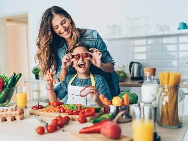 Uma mãe e sua criança estão na cozinha, com diversos alimentos sobre uma bancada. Ela segura dois tomates pequenos na altura dos olhos da criança e ambas estão sorrindo Uma mãe e sua criança estão na cozinha, com diversos alimentos sobre uma bancada. Ela segura dois tomates pequenos na altura dos olhos da criança e ambas estão sorrindo