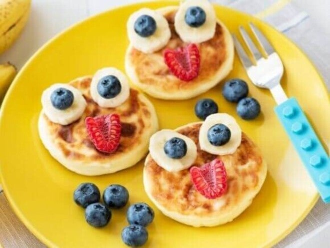 Três panquecas douradas decoradas de forma lúdica com fatias de banana, mirtilos e framboesas, formando rostos divertidos.