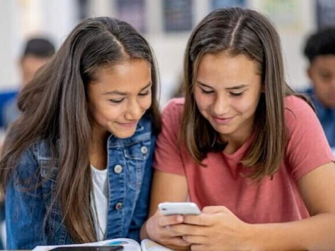 Duas meninas sentadas uma ao lado da outra na sala de aula, sorrindo e olhando para um celular Duas meninas sentadas uma ao lado da outra na sala de aula, sorrindo e olhando para um celular