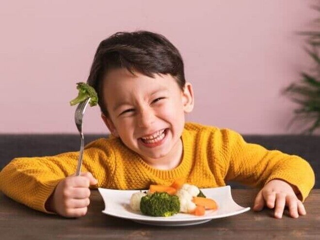 Menino pequeno sorrindo com entusiasmo ao segurar um garfo com brócolis. À sua frente, um prato com brócolis, cenoura e couve-flor ilustra como montar um prato saudável infantil de forma atrativa e nutritiva para crianças.