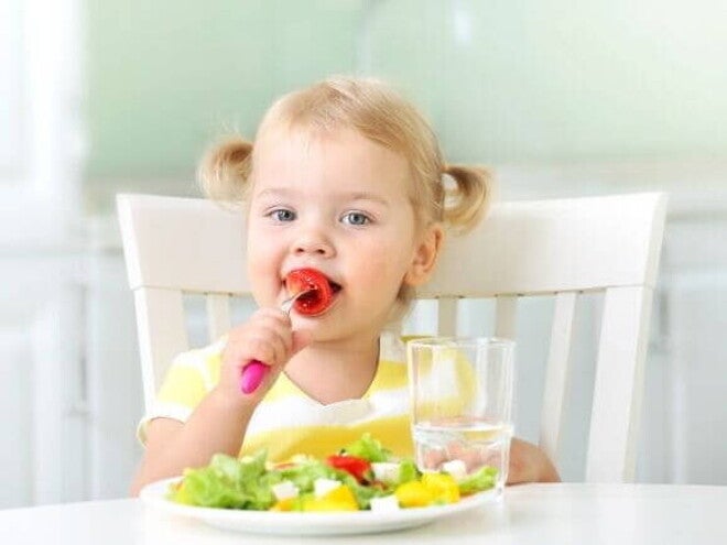 Criança pequena sentada à mesa, sorrindo e comendo salada em um prato, com um copo d'água ao lado.