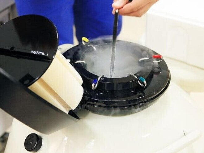 Close-up de um tanque de nitrogênio líquido aberto, com vapor frio subindo. Uma pessoa está inserindo ou retirando uma palheta ou recipiente de armazenamento de embriões.