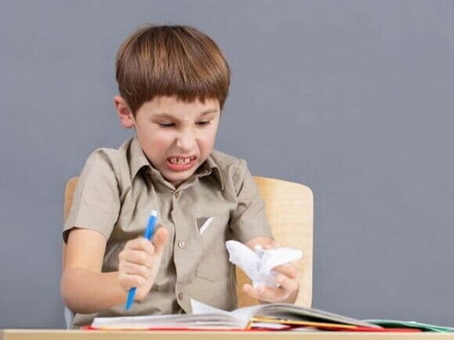 Menino em idade escolar sentado em uma carteira, com expressão de raiva ou frustração, cerrando os dentes e segurando um lápis ou caneta com força.