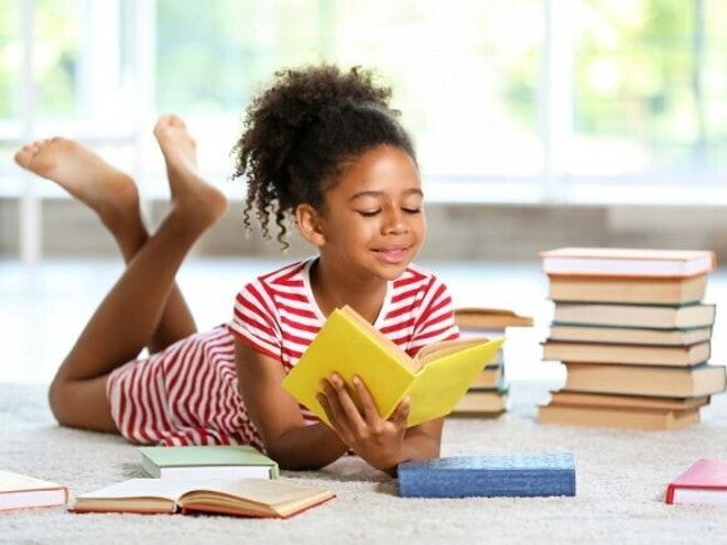 Uma menina sorridente, está deitada de bruços em um tapete, lendo um livro. Vários outros livros estão espalhados ao redor dela.