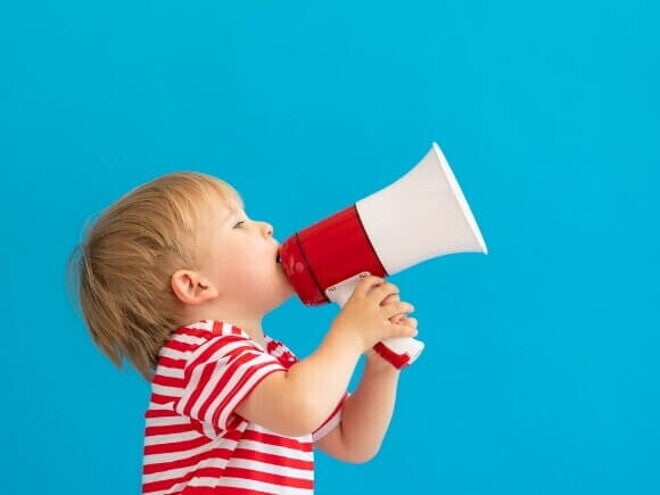 Um menino loiro usando camiseta listrada segura um megafone vermelho e branco contra um fundo azul sólido.